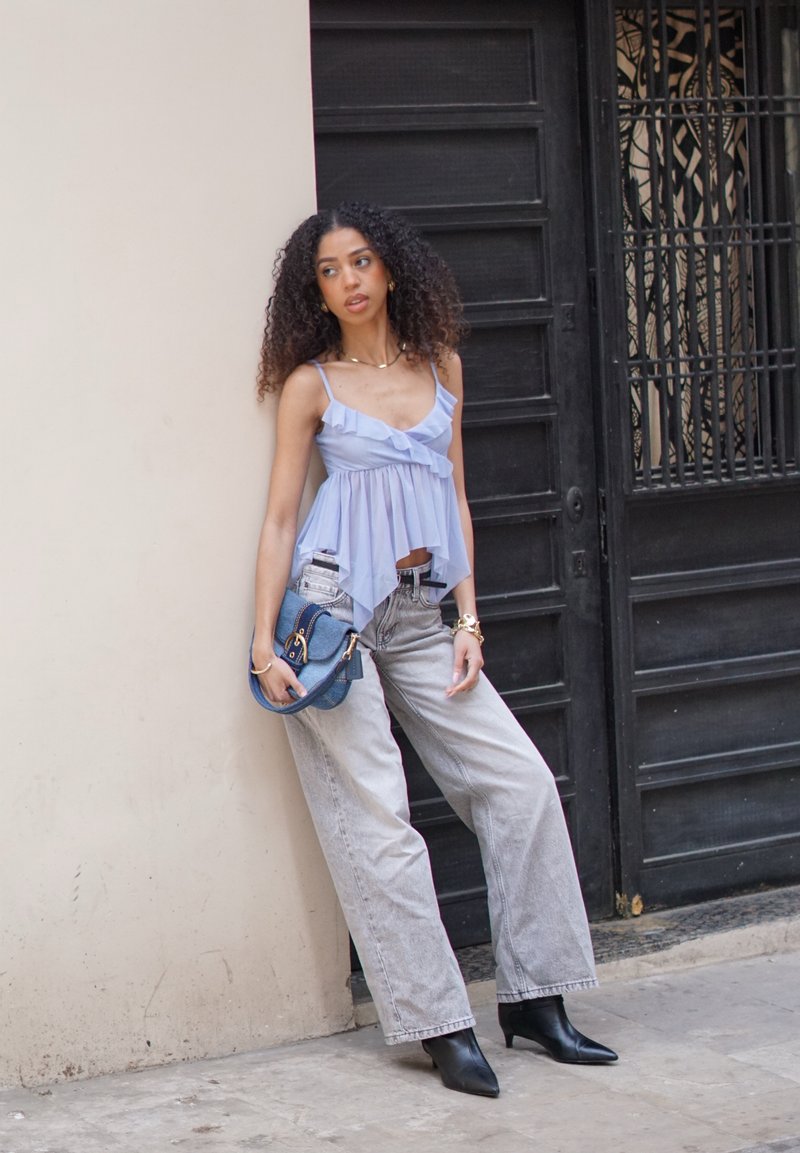 Femme aux cheveux bouclés portant un haut sans manches bleu clair et un jean large gris, penchée contre un mur beige près d'une porte noire, tenant un sac bleu.