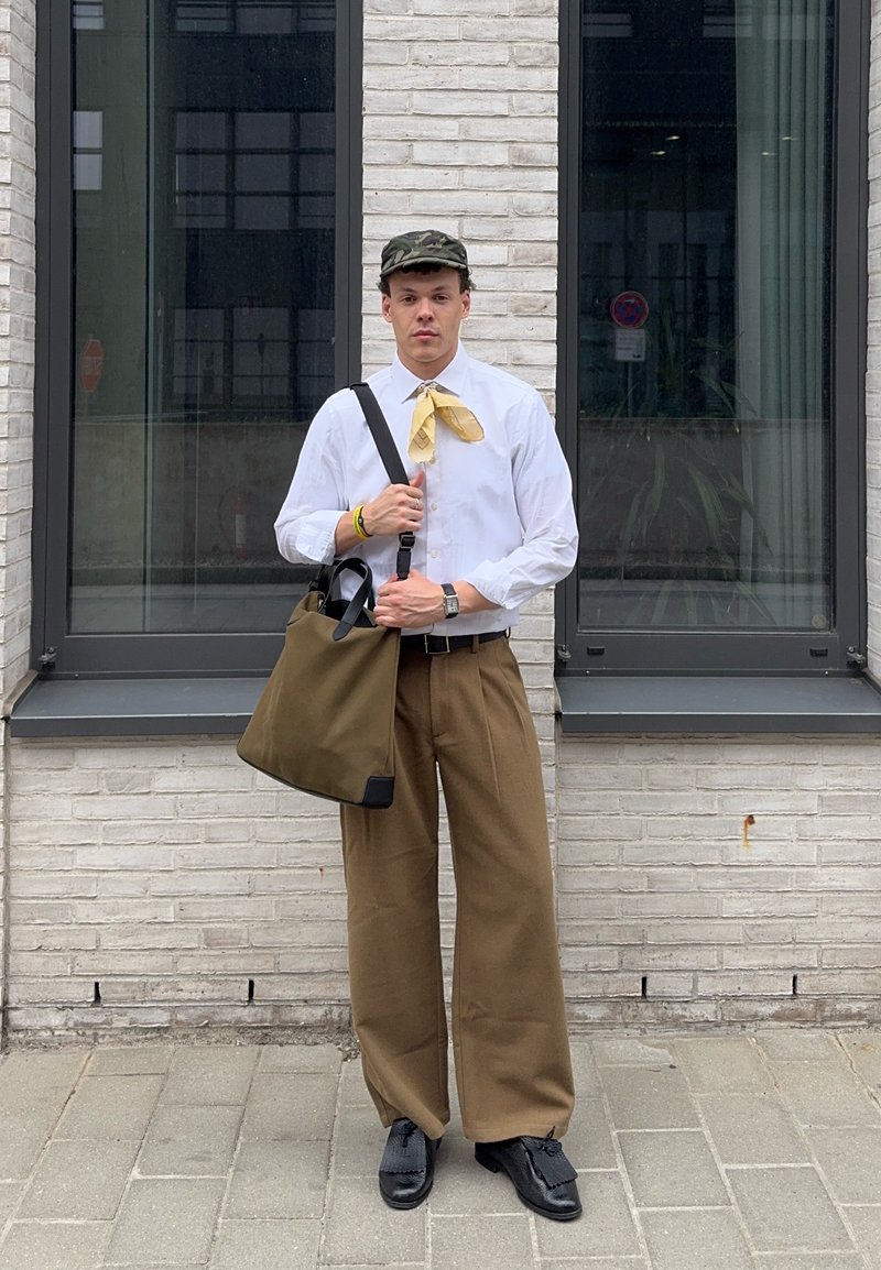 Young man wearing white shirt, brown wide trousers, yellow neck scarf, camo cap, black shoes, holding green tote bag, standing by a white brick wall.