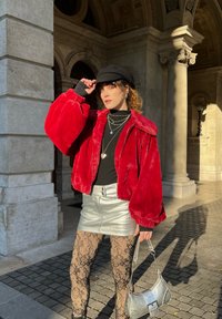 Rød faux fur bomberjakke, svart topp, sølvmetallisk miniskjørt, svarte blonde strømpebukser, sølvhåndveske, solfylt steinarkitektur som bakgrunn.