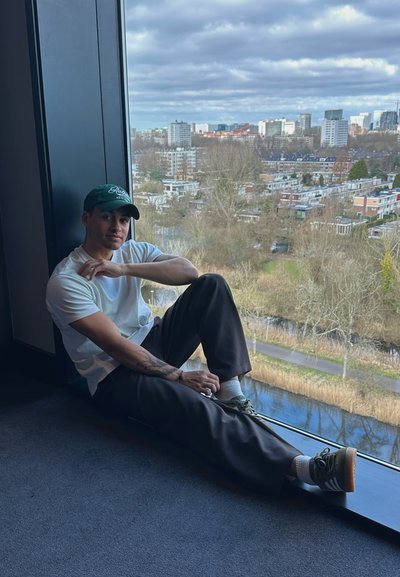 Una persona con una camiseta blanca y pantalones oscuros se sienta en el alféizar de una ventana, mirando un paisaje urbano. Lleva una gorra verde y zapatos con acentos verdes.