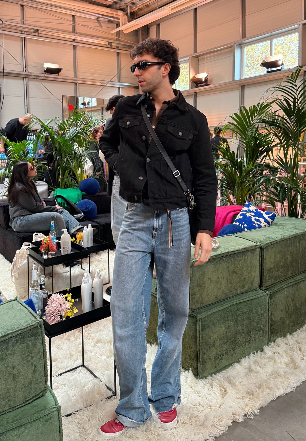 Giacca di jeans nera, sbottonata sopra una camicia marrone, abbinata a jeans blu larghi e scarpe rosse. Tiene una borsa nera, circondato da vegetazione.