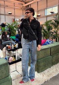 Giacca di jeans nera, sbottonata sopra una camicia marrone, abbinata a jeans blu larghi e scarpe rosse. Tiene una borsa nera, circondato da vegetazione.