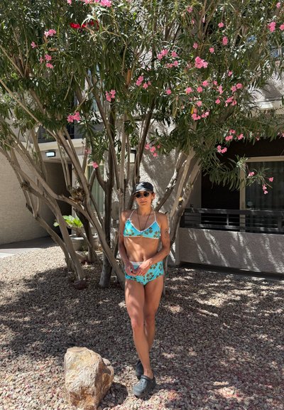 Mujer con bikini azul con estampado floral, gafas de sol y gorra negra, de pie sobre terreno rocoso bajo un árbol en flor junto al balcón de un edificio.