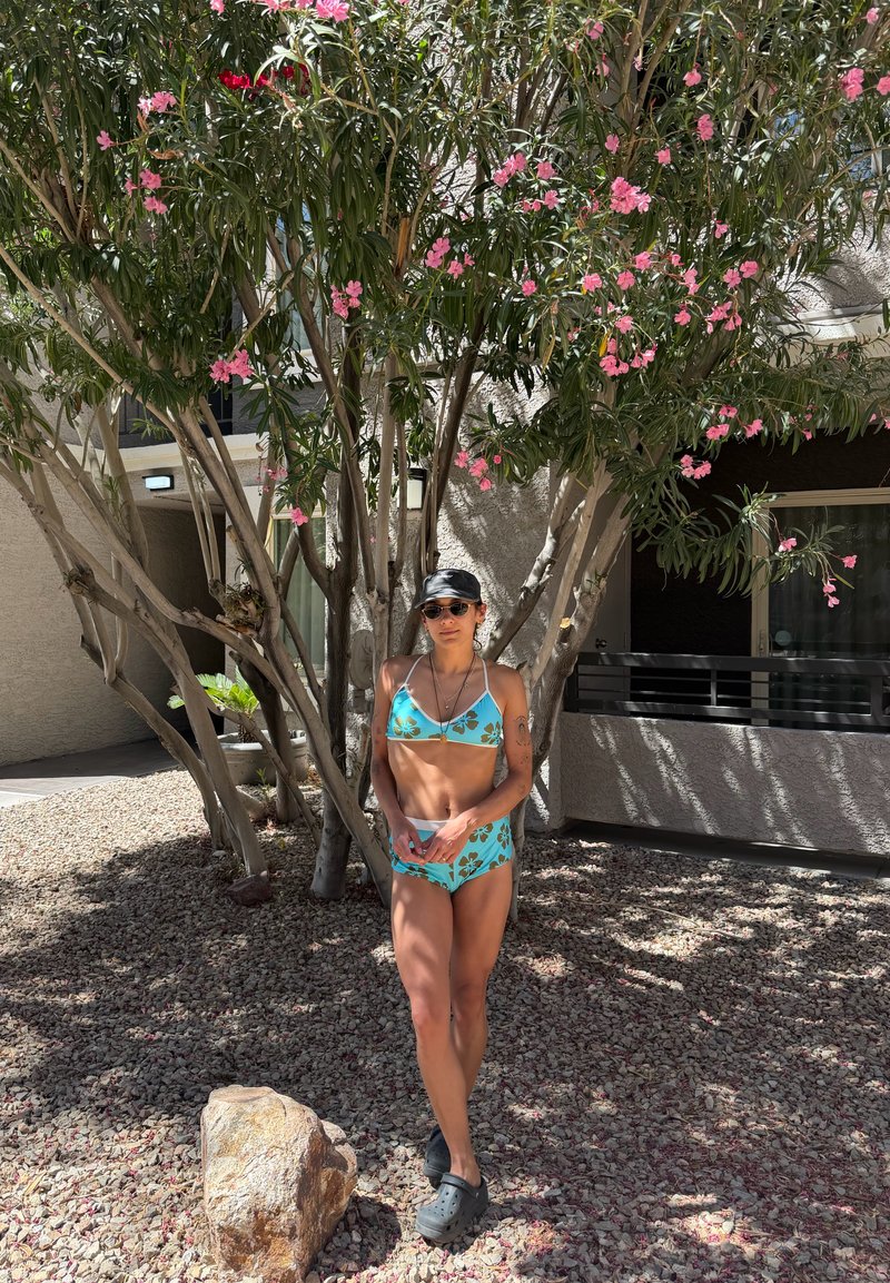 Femme portant un bikini bleu à motifs floraux, des lunettes de soleil et une casquette noire, debout sur un sol rocheux sous un arbre en fleurs près d'un balcon d'immeuble.