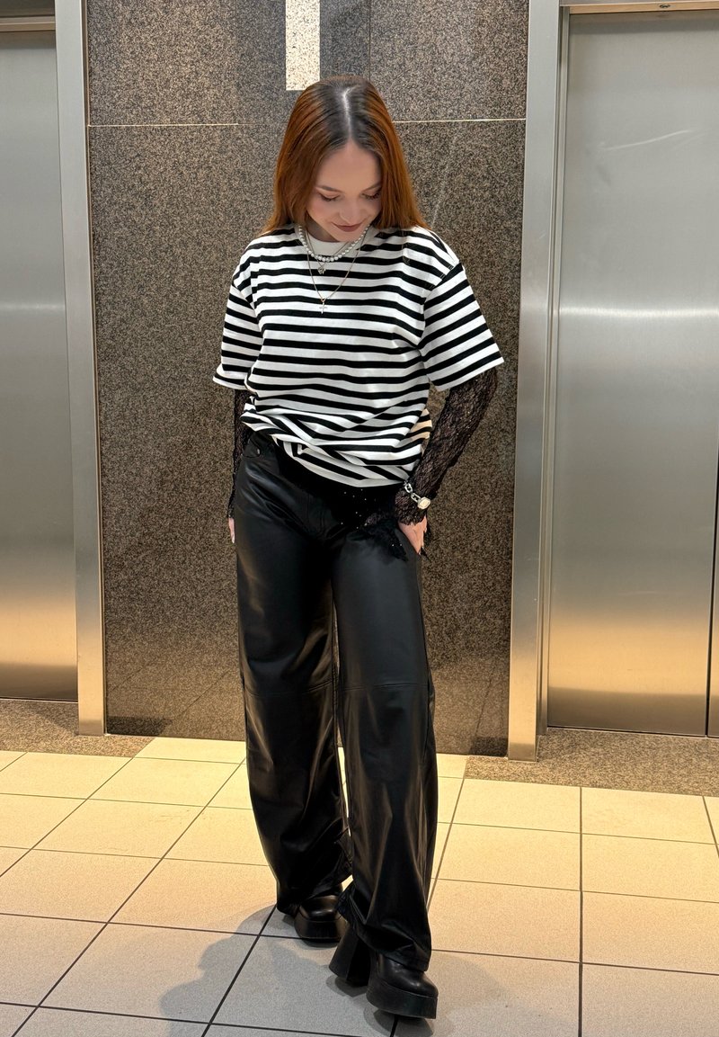 Femme portant un pantalon en cuir noir, des manches en dentelle noire sous un t-shirt rayé noir et blanc, debout entre deux ascenseurs à l'intérieur.