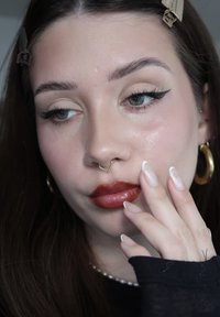 Jeune femme avec un eye-liner ailé, un rouge à lèvres rouge foncé, un anneau de septum doré, un grand anneau créole et une manucure française, touchant son visage avec les cheveux attachés.