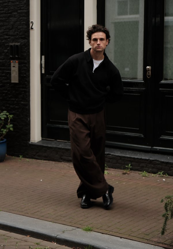 Ein Mann, der einen schwarzen Pullover und braune weite Hosen trägt, steht mit den Händen hinter dem Rücken auf einem gepflasterten Gehweg vor einer schwarzen Tür.