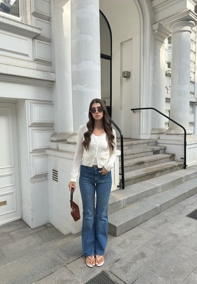 Mujer con gafas de sol, cárdigan blanco, vaqueros azules, sosteniendo un bolso marrón, de pie frente a un edificio blanco con columnas y escaleras.