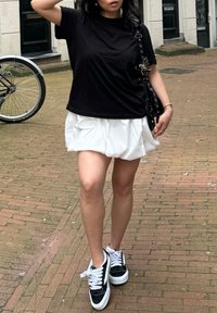 T-shirt noir à manches courtes, jupe blanche à couches, baskets noir et blanc avec lacets. L'arrière-plan comporte un vélo et un pavé en briques.