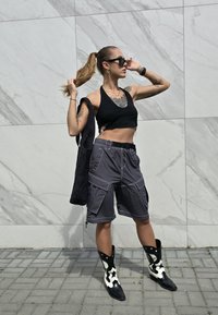 Μαύρο τοπ με λαιμόκοψη, γκρι cargo σορτς με τσέπες, μαύρες και λευκές μοτίβο μπότες μέχρι το γόνατο, και μαύρη τσάντα ώμου μπροστά σε μαρμαροστρωτό τοίχο.