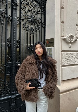 Joven mujer con cabello largo y negro, que lleva un abrigo marrón de pelo y pantalones blancos, sosteniendo un bolso negro, de pie junto a una puerta de hierro negro decorativa.