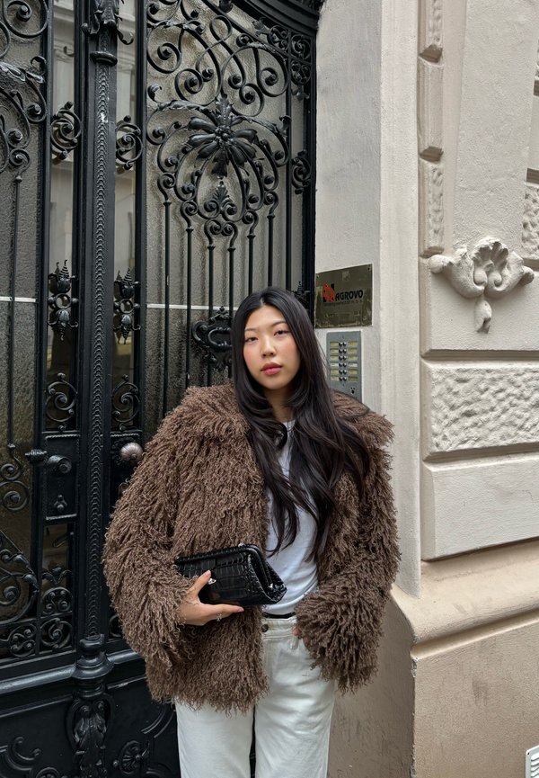 Młoda kobieta z długimi czarnymi włosami, ubrana w brązowy futrzany płaszcz i białe spodnie, trzymająca czarną kopertówkę, stojąca przy ozdobnej czarnej żelaznej bramie.