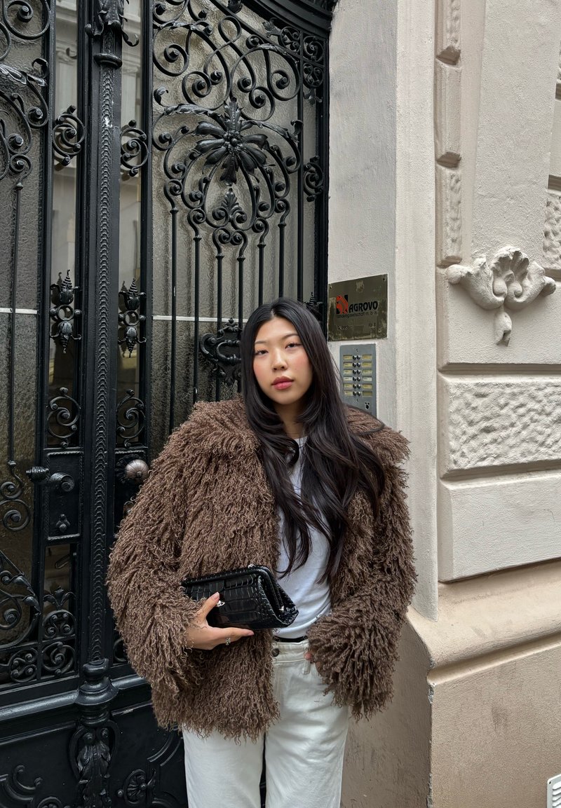 Jeune femme aux longs cheveux noirs portant un manteau marron en fourrure et un pantalon blanc, tenant une pochette noire, debout près d'un portail en fer noir décoratif.