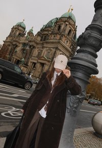 Barna kordbársonykabát, barna mellény és fehér ing fölött rétegezve, kiegészítve egy világos bézs sapkával logóval, fekete táskával és sötét szürke nadrággal.