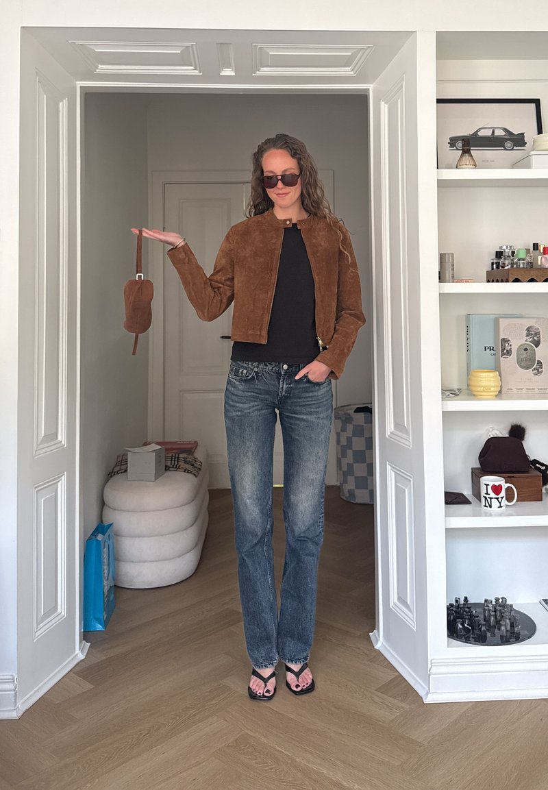 Femme en veste en daim marron, haut noir, jean bleu et sandales, debout dans l’encadrement de la porte tenant un petit sac marron avec des lunettes de soleil.