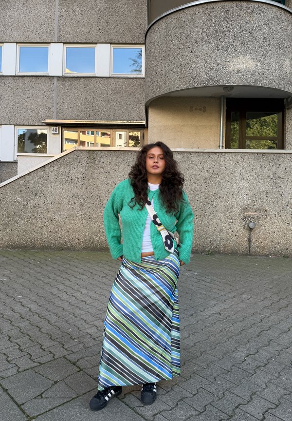 Une femme se tient devant un immeuble d’habitation en béton. Elle porte un cardigan vert, une jupe à rayures et des baskets, ainsi qu’un sac en bandoulière.