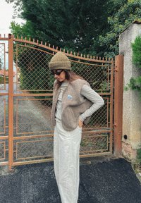 Jasnobrązowa kamizelka z polaru na szarym długim rękawie, w połączeniu z kremowymi spodniami o szerokich nogawkach. Zielonooliwkowa wełniana czapka i okulary przeciwsłoneczne.