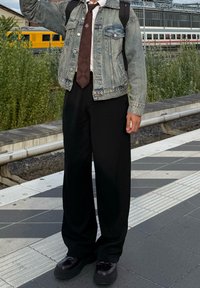 Osoba oblečená v džínsovej bunde, bordovej viazanke so vzorom, čiernych nohaviciach a čiernych topánkach stojí na nástupišti železničnej stanice vedľa koľají.