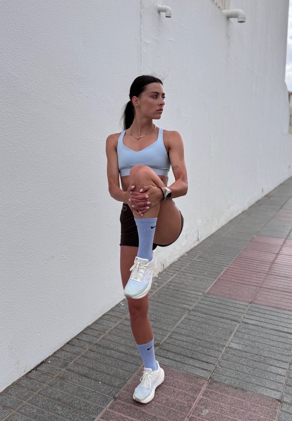 Svetlo modra športna modrc, rjave kratke hlače, svetlo modre Nike nogavice in bele športne čevlje. V stoječem položaju z eno koleno dvignjenim, ob beli steni.