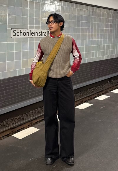 Chaleco beige de punto de gran tamaño sobre una camisa roja de manga larga, pantalones negros de pierna ancha y un bolso cruzado color caqui en una estación de metro.