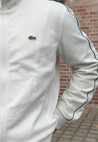 Pessoa vestindo uma jaqueta branca com fecho éclair e detalhe preto nas mangas, com um pequeno logotipo de crocodilo verde no peito, em pé ao ar livre perto de tijolos.