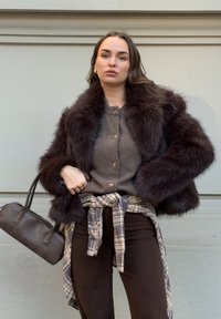 Femme portant une veste en fausse fourrure marron foncé, un pull gris boutonné, une chemise à carreaux nouée à la taille, un pantalon foncé, tenant un sac à main marron contre un mur.