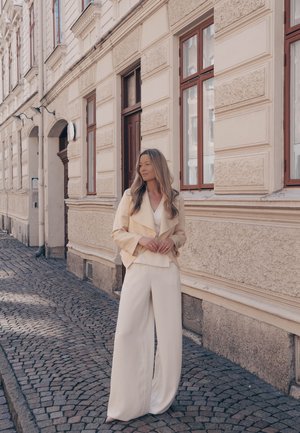 Mujer con blazer crema y pantalones anchos de pie en una calle empedrada junto a un edificio beige con textura y ventanas altas.