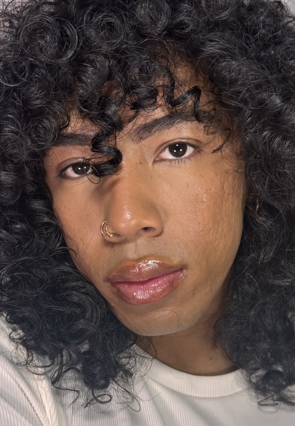 Lähikuva kiharat mustat hiukset omaavasta kasvosta. Kasvoilla on kultainen rengas nenässä, sileä iho ja kiiltävät huulet. Valkoinen teksturoitu paita on näkyvissä.