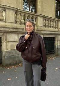 Femme en veste en cuir marron et pantalon gris debout dans une rue de la ville, tenant un sac à main marron texturé, ajustant son col près d'une balustrade en pierre.