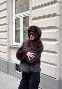 Jachetă din blană artificială maro închis, cu o pălărie asortată, având o textură pufoasă și mâneci strâmte, purtată peste pantaloni negri.