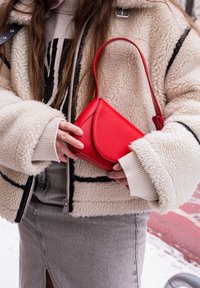 Άτομο με μπεζ μπουφάν shearling και γκρι φούστα κρατά μια μικρή κόκκινη δερμάτινη τσάντα χειρός με πάνω λαβή σε εξωτερικό χώρο.