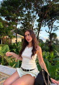 Valkoinen hihaton paita, jossa liehuva, laskostettu vihreä hame, musta vyö ja musta käsilaukku. Taustalla rehevää vihreää kasvillisuutta ja puita.