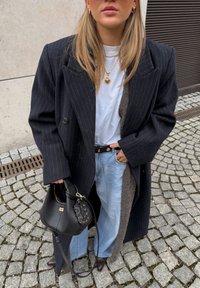 Manteau oversize noir à fines rayures, t-shirt blanc, jean bleu clair ample, sac à main noir avec des accents dorés, et chaussures marron à bout pointu.