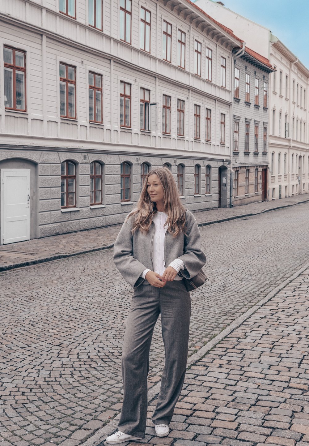Grå ullblandet dress med vide bukser, kombinert med en hvit teksturert genser. Bakgrunnen viser brosteinsbelagte gater og historiske bygninger.
