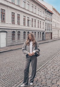 Grå ullblandet dress med vide bukser, kombinert med en hvit teksturert genser. Bakgrunnen viser brosteinsbelagte gater og historiske bygninger.