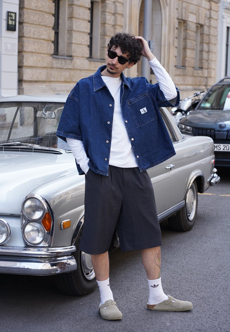 Τζάκετ denim, λευκό t-shirt, μαύρα σορτς, μπεζ παντόφλες και γυαλιά ηλίου. Στηρίζει κοντά σε ένα vintage ασημί αυτοκίνητο σε δρόμο της πόλης.