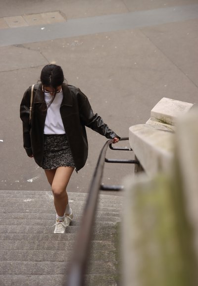 Persona subiendo escaleras de piedra, vistiendo una chaqueta de cuero oscura, una camisa blanca, una falda negra texturizada y zapatillas blancas con calcetines estampados.
