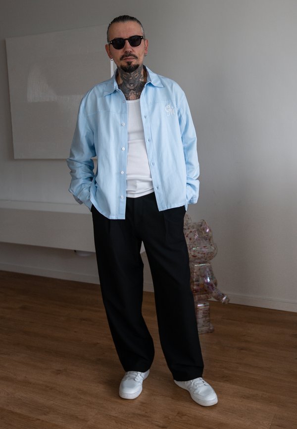 Homme avec des tatouages et des lunettes de soleil, les mains dans les poches, portant une chemise bleu clair, un débardeur blanc, un pantalon noir et des baskets blanches à l’intérieur.