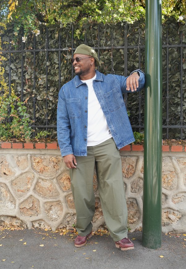 Un hombre sonriente con gafas de sol y un gorro verde se apoya en un poste verde junto a una verja de hierro cubierta de hojas.