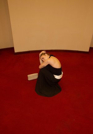 Mujer con un vestido negro sin espalda sentada acurrucada sobre una alfombra roja, apoyando la cabeza en la rodilla, con un pequeño bolso beige tejido cerca.