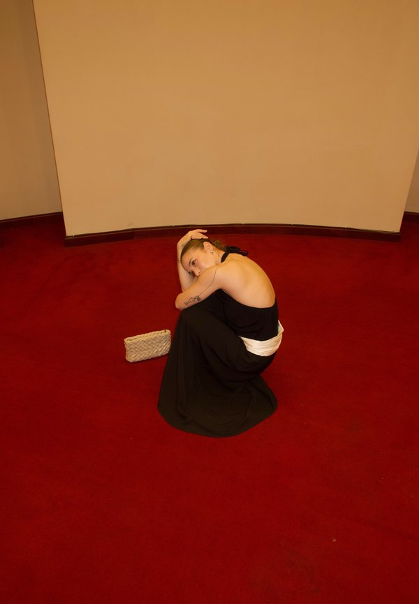 Ženska v črni obleki brez hrbta sedi skrčena na rdeči preprogi, glavo naslonjeno na koleno, ob njej majhna bež pletena torbica.