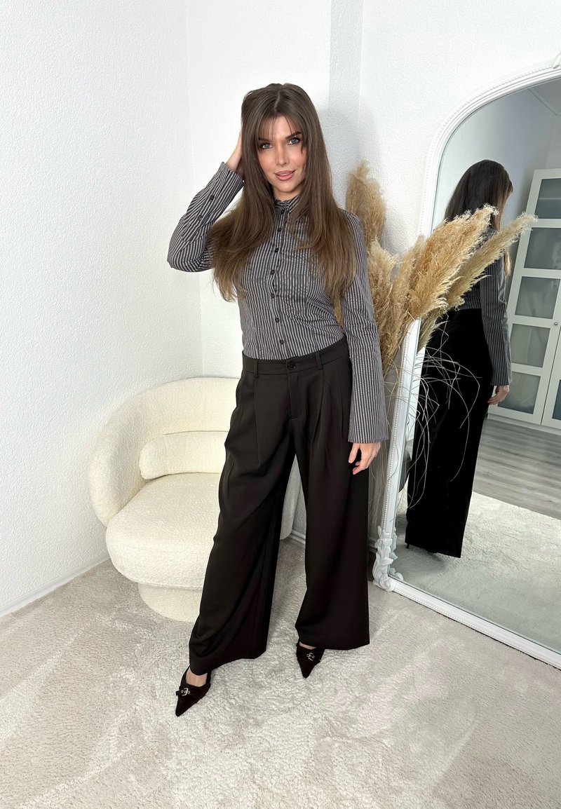 Woman with long brown hair wearing a striped button-up shirt and wide black trousers stands by a round white chair and tall mirror with dried plants.