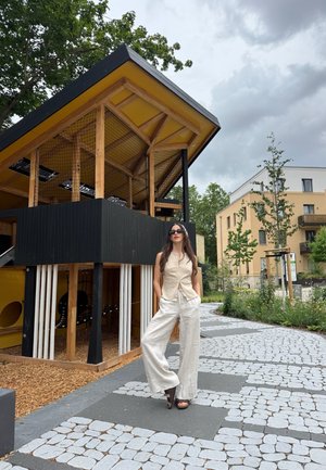 Mujer con gafas de sol y ropa ligera está de pie en un camino de piedra con patrón cerca de una estructura moderna de madera para juegos y edificios residenciales.