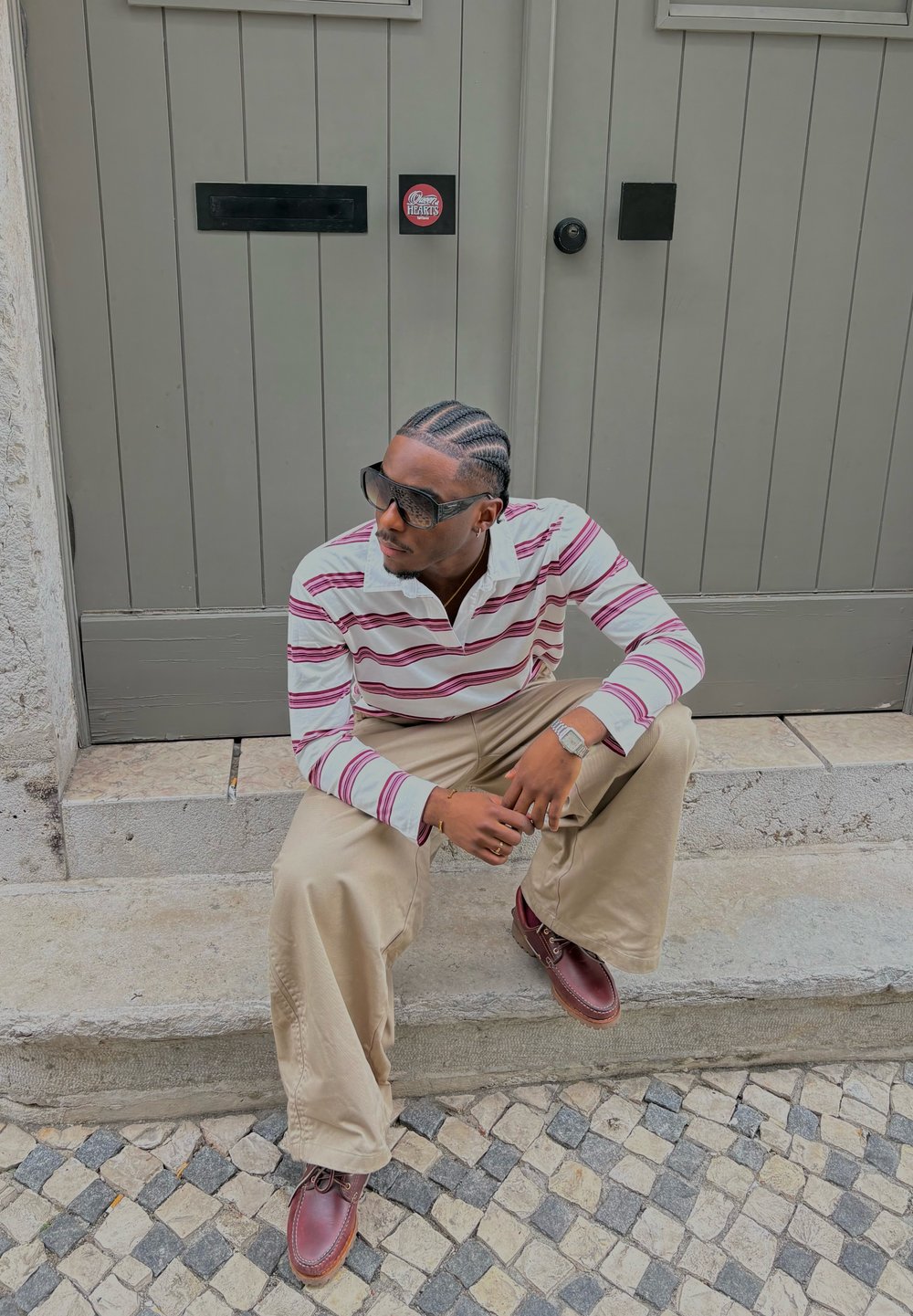 Polo à manches longues rayé avec des couleurs blanche et bordeaux, pantalon large beige, et chaussures à lacets marron, assis sur des marches en pierre.