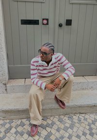 Polo à manches longues rayé avec des couleurs blanche et bordeaux, pantalon large beige, et chaussures à lacets marron, assis sur des marches en pierre.