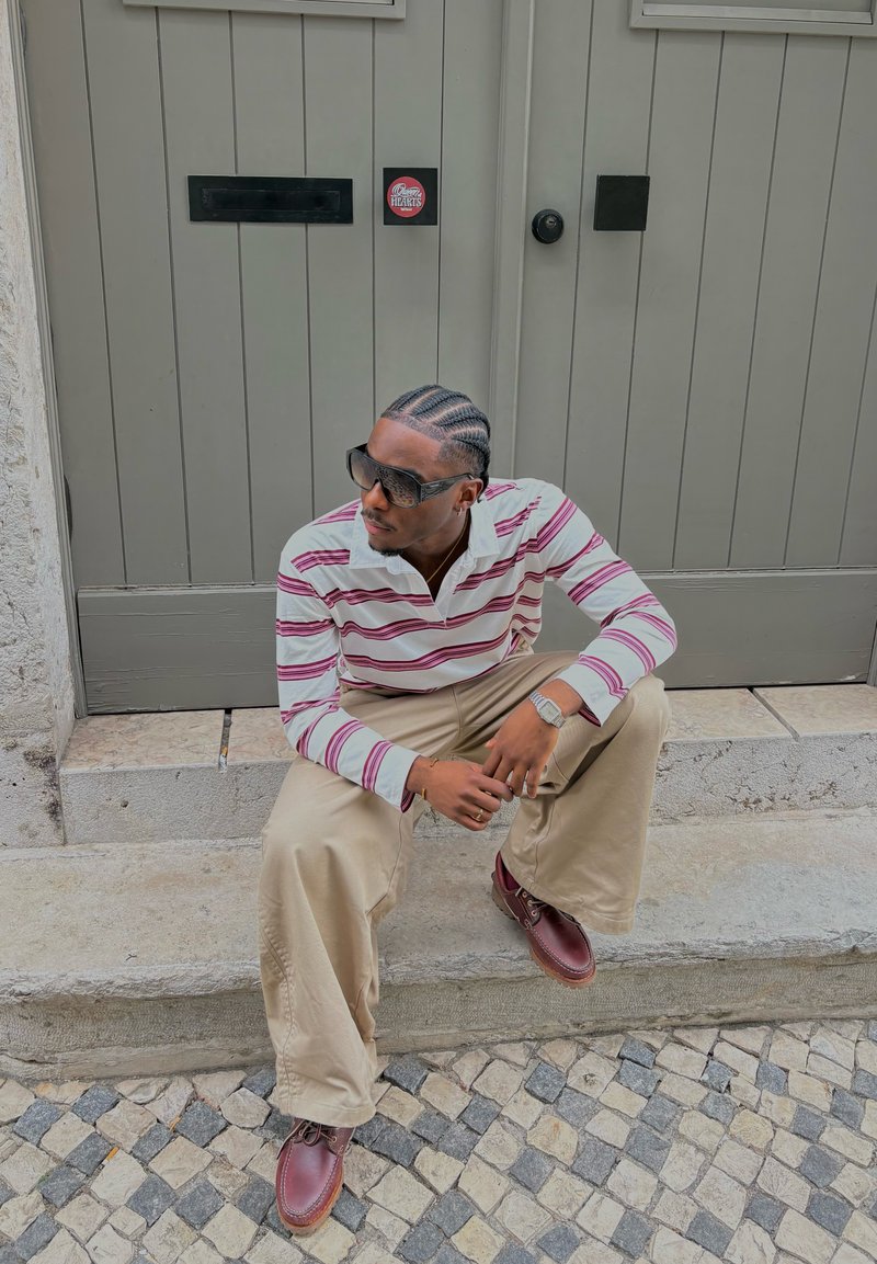 Ribaga pikiga polo-särg pikki varrukatega valgete ja burgundia värvitoonidega, beežid laiad püksid ja pruunid nööbiga kingad, istub kivist treppidel.