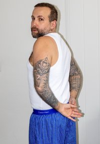Chemise blanche sans manches et caleçons à carreaux bleus avec une ceinture Calvin Klein ; plusieurs tatouages sur le bras ; debout de profil.