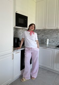 T-shirt bianco, pantaloni da pigiama a fantasia rosa con coulisse e sandali beige; in piedi vicino a un piano di marmo in una cucina moderna.