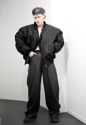 Persona con chaqueta bomber negra de gran tamaño, pantalones de cuero de pierna ancha, boina negra y zapatos negros, de pie contra una pared blanca en la esquina.