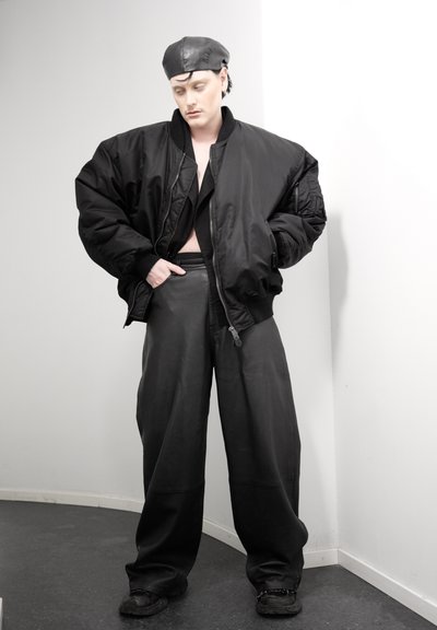 Persona con chaqueta bomber negra de gran tamaño, pantalones de cuero de pierna ancha, boina negra y zapatos negros, de pie contra una pared blanca en la esquina.
